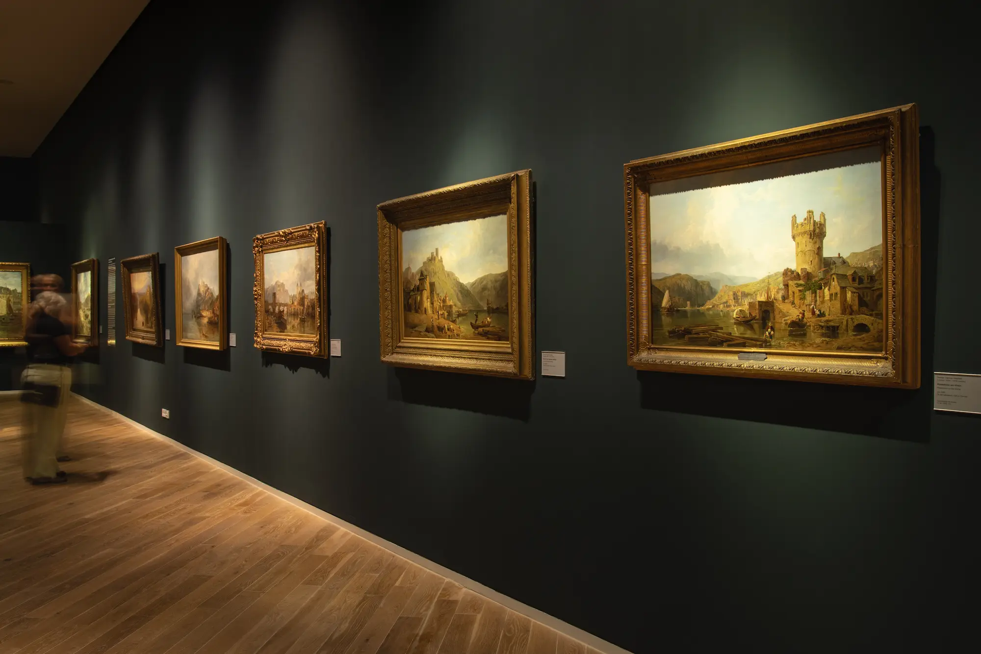 Innenansicht einer Bildergalerie im Museum (vermutlich Mittelrhein Museum Koblenz) mit traditionellen Ölgemälden in goldfarbenen Rahmen an einer dunklen Wand und einem Besucher im Unschärfebereich.