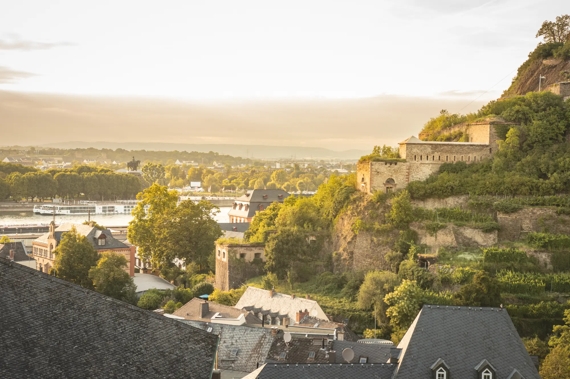 Historische Mauern der Festung Ehrenbreitstein im Vordergrund, mit Blick über die Dächer der Altstadt auf den Zusammenfluss von Rhein und Mosel (Deutsches Eck) in Koblenz.