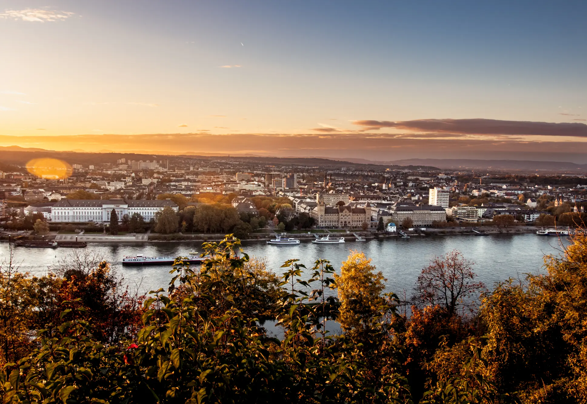 Panoramablick auf die Stadt Koblenz und den Rhein in warmem Herbstlicht, gesehen von einem erhöhten Aussichtspunkt mit bunten Bäumen im Vordergrund.