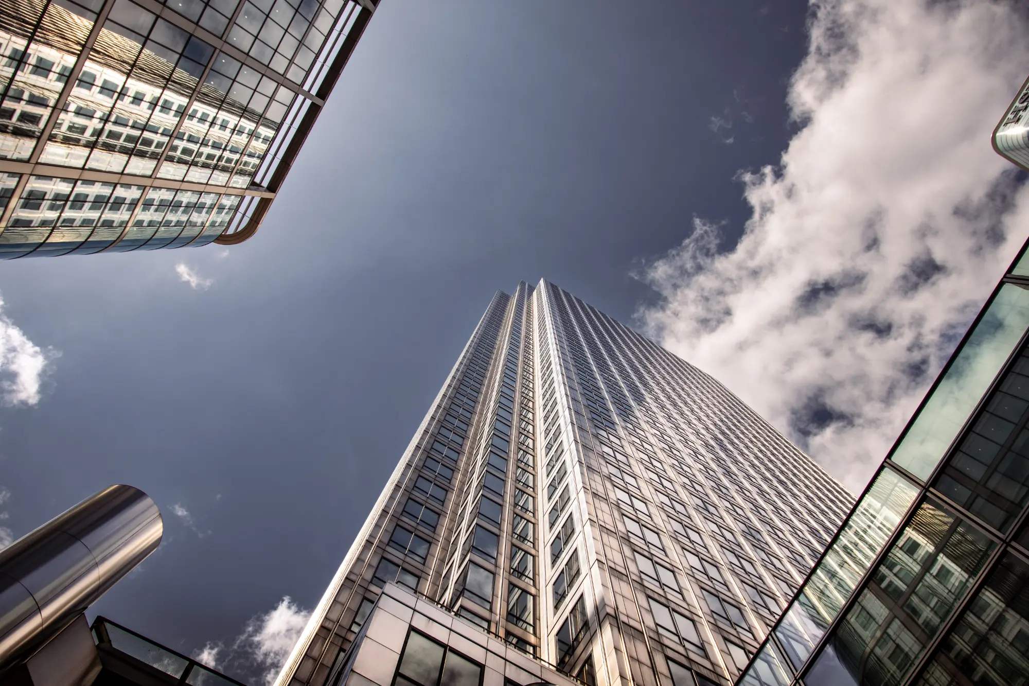 Perspektive von unten auf den One Canada Square Wolkenkratzer in London Canary Wharf, umrahmt von reflektierenden Glasfassaden anderer Hochhäuser und blauem Himmel.