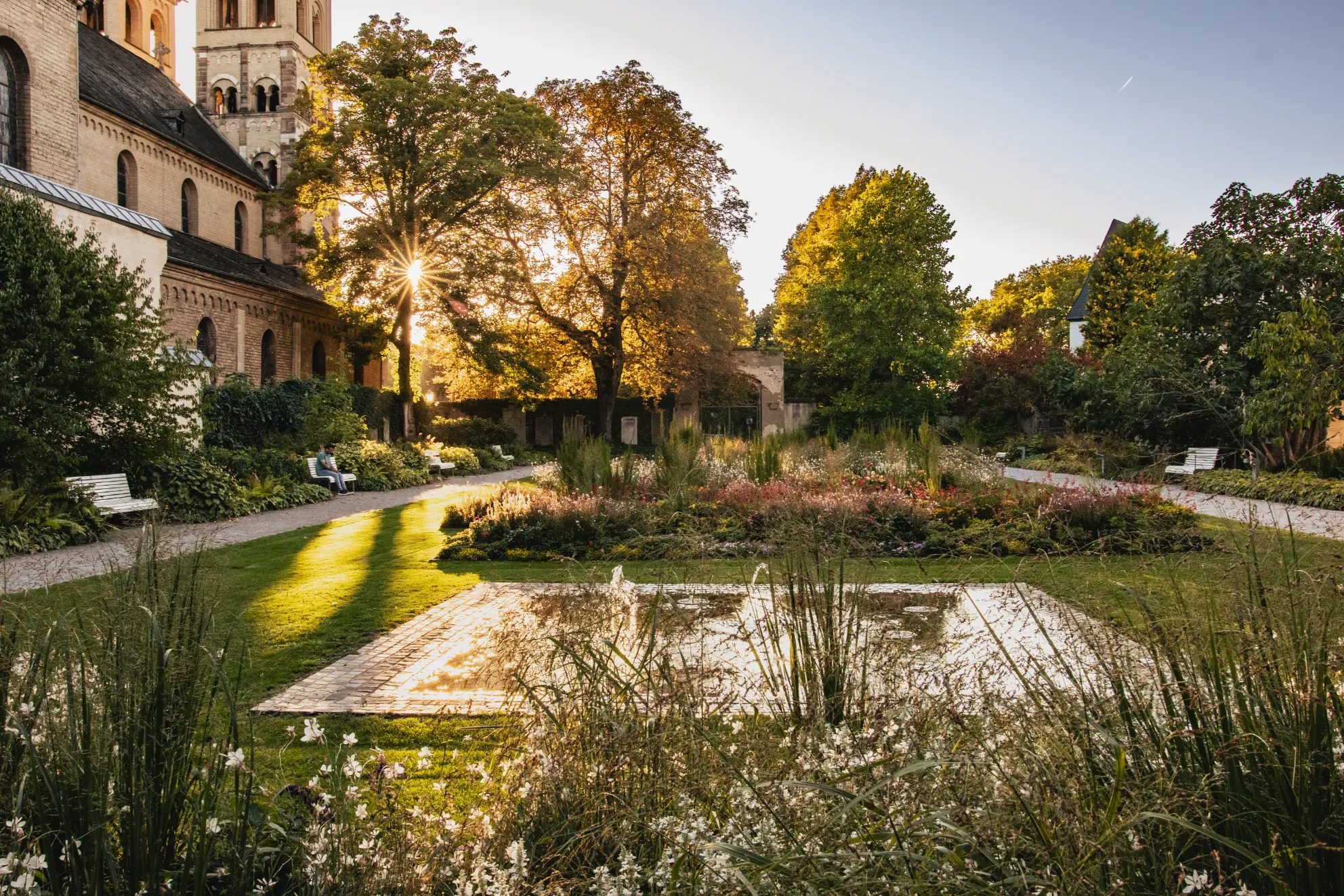 Der blühende Blumenhof in Koblenz (Kastorhof) in warmem Abendlicht, mit einem kleinen Wasserbecken, Blumenbeeten und der romanischen Basilika St. Kastor im Hintergrund.