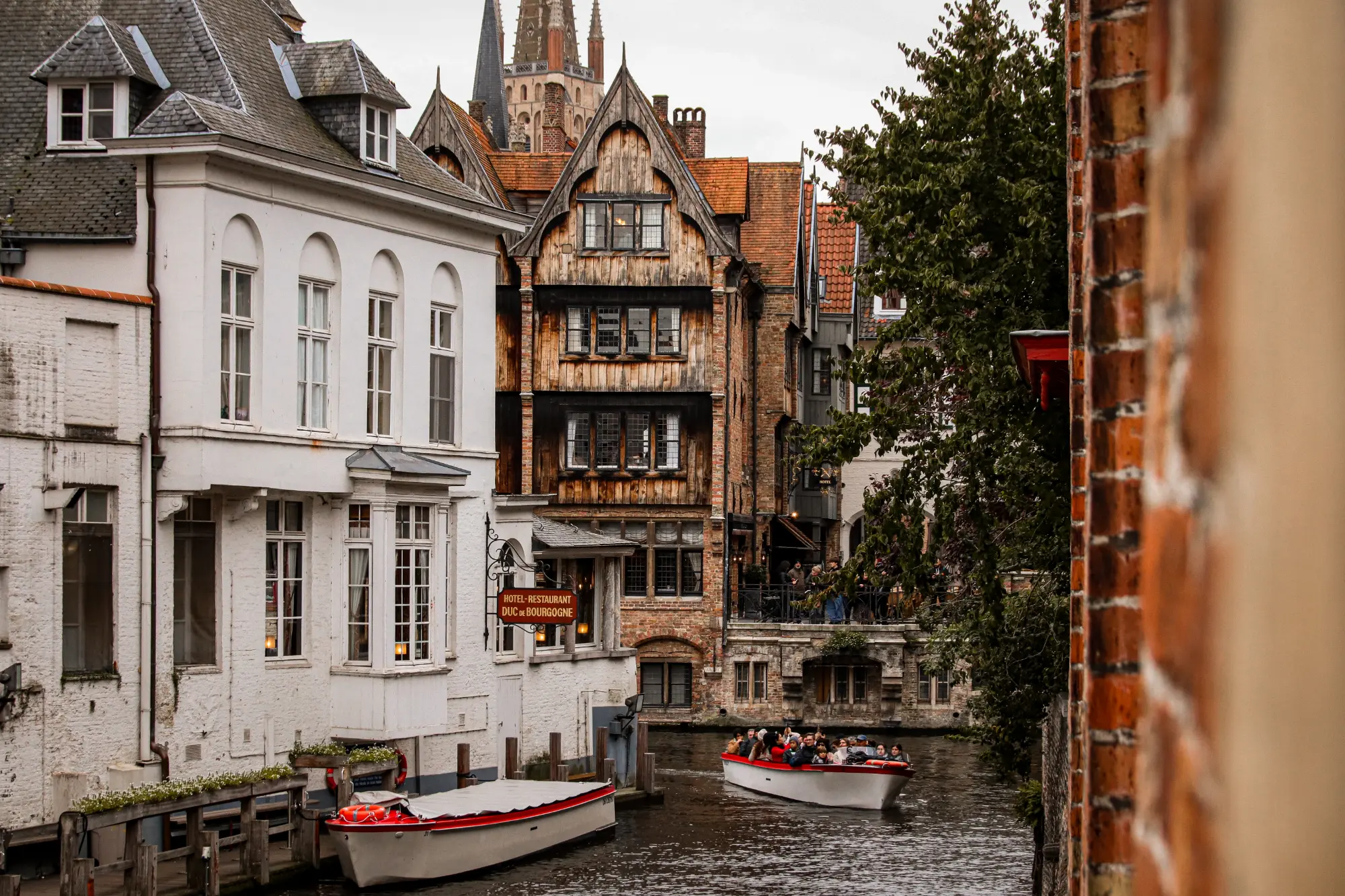 Ansicht historischer Häuser am Kanal in der Altstadt von Brügge (Belgien) an einem grauen Tag, mit einem Touristenschiff auf dem Wasser und einem Glockenturm im Hintergrund.