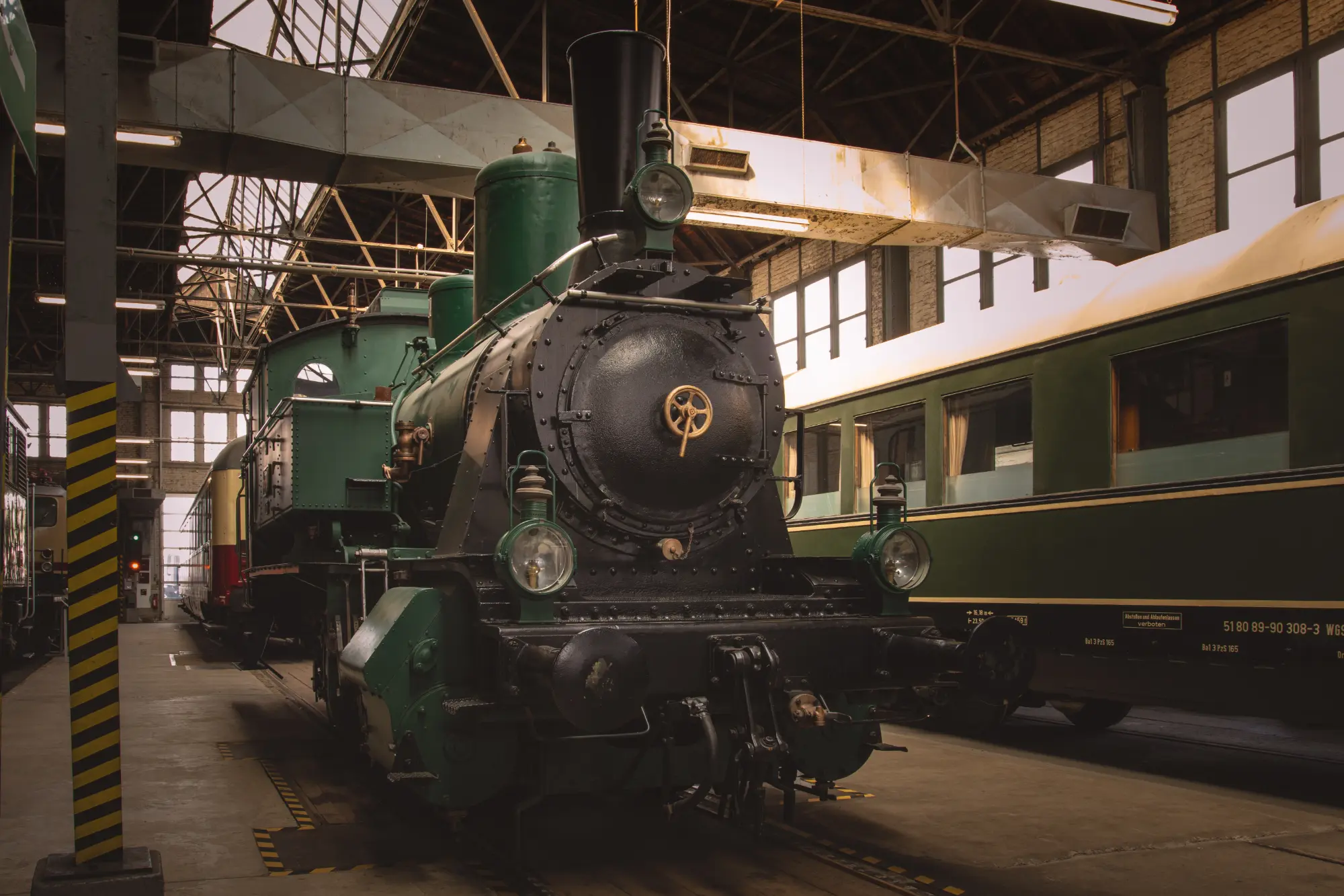 Frontalansicht einer historischen, dunkelgrünen Dampflokomotive in einer Halle des DB Museums (vermutlich Koblenz-Lützel), umgeben von anderen historischen Waggons.