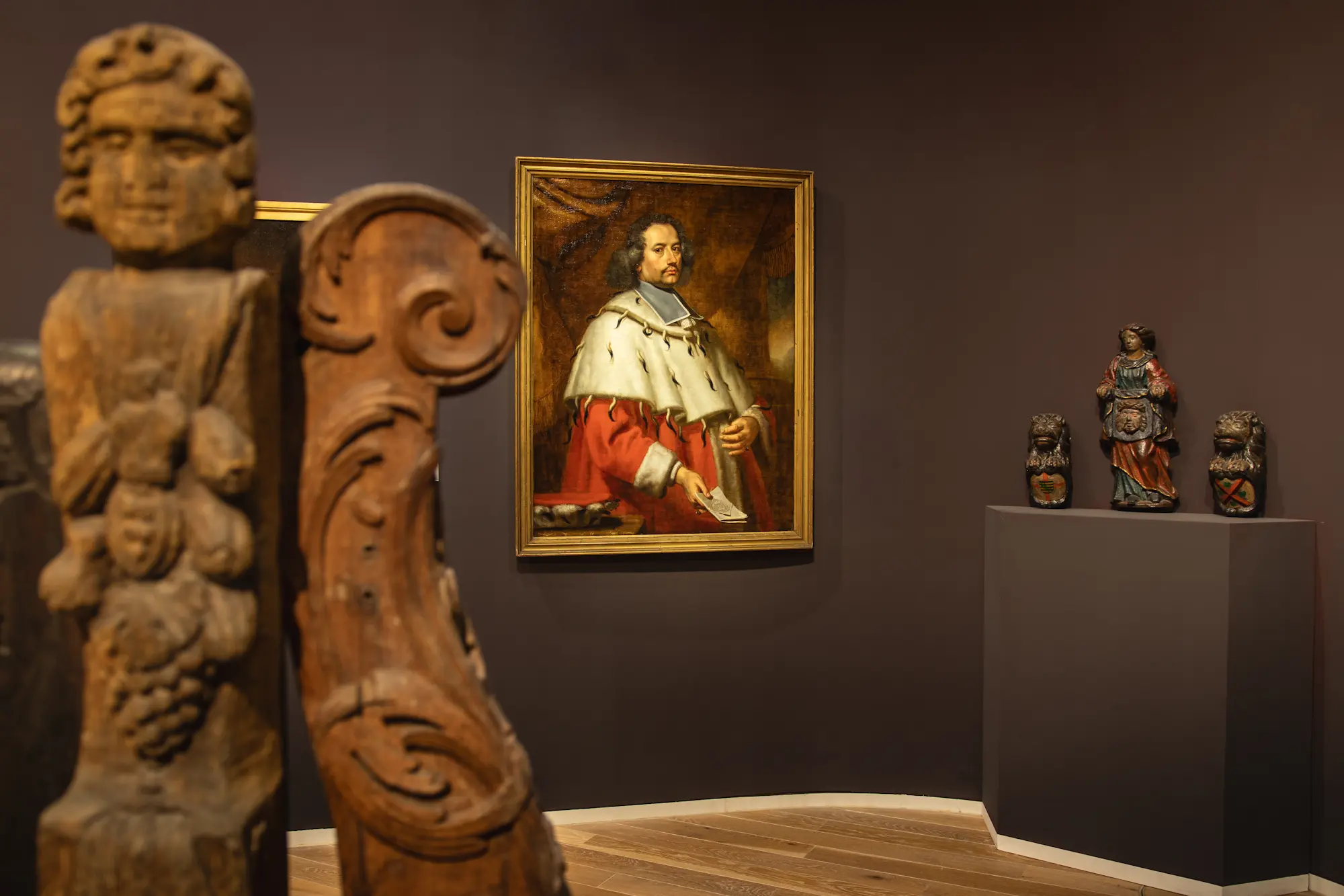 Innenansicht des Mittelrhein Museums in Koblenz: Eine moderne Ausstellung mit einer hölzernen Skulptur im Vordergrund und einem barocken Porträtgemälde eines Kurfürsten oder Adligen im Hintergrund.