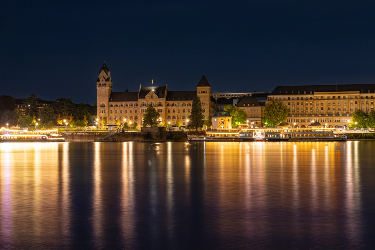 Historische Gebäude, darunter das ehemalige Regierungsgbäude in Koblenz, beleuchtet bei Nacht am Rheinufer mit Spiegelungen im Wasser.