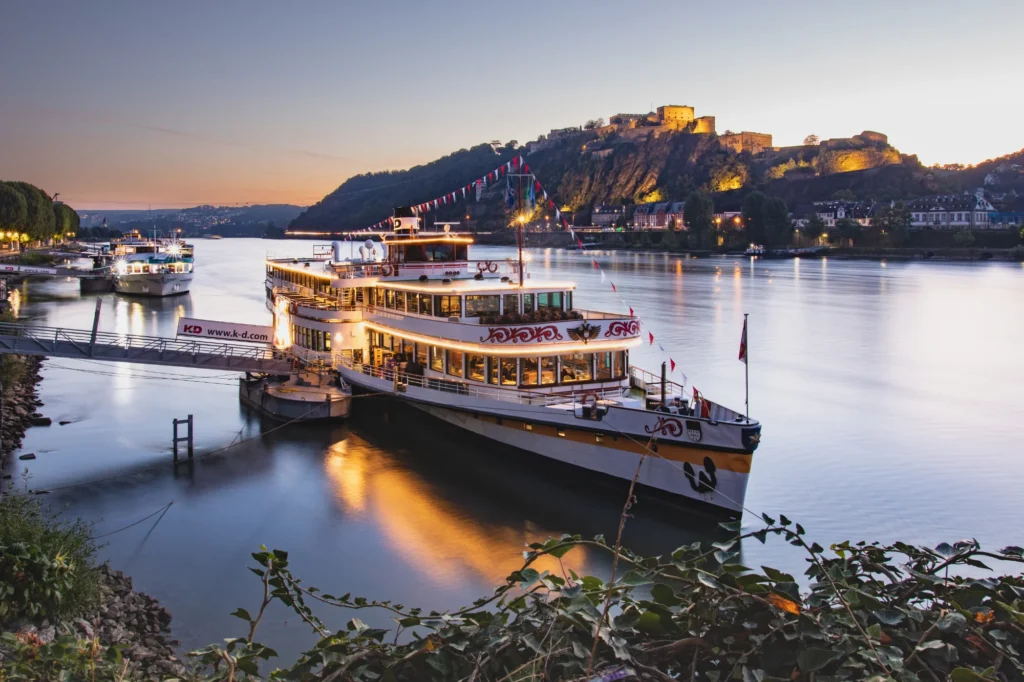 Beleuchtetes KD-Flusskreuzfahrtschiff legt im Morgengrauen am Steg an, mit der erleuchteten Festung Ehrenbreitstein auf dem Hügel im Hintergrund in Koblenz.