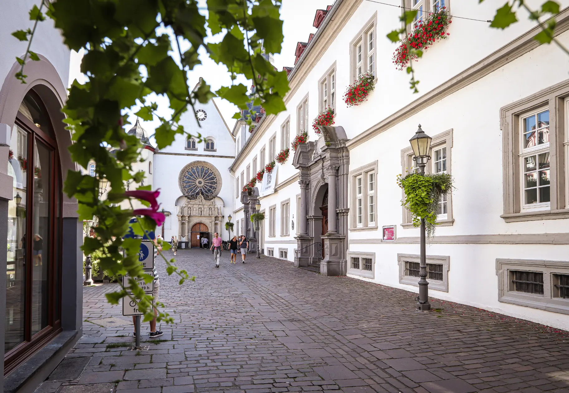 Der Jesuitenplatz in Koblenz mit dem hellen, langen Barockgebäude (ehemaliges Jesuitenkolleg) und dem romanischen Portal der Citykirche im Hintergrund, eingerahmt von Laub.