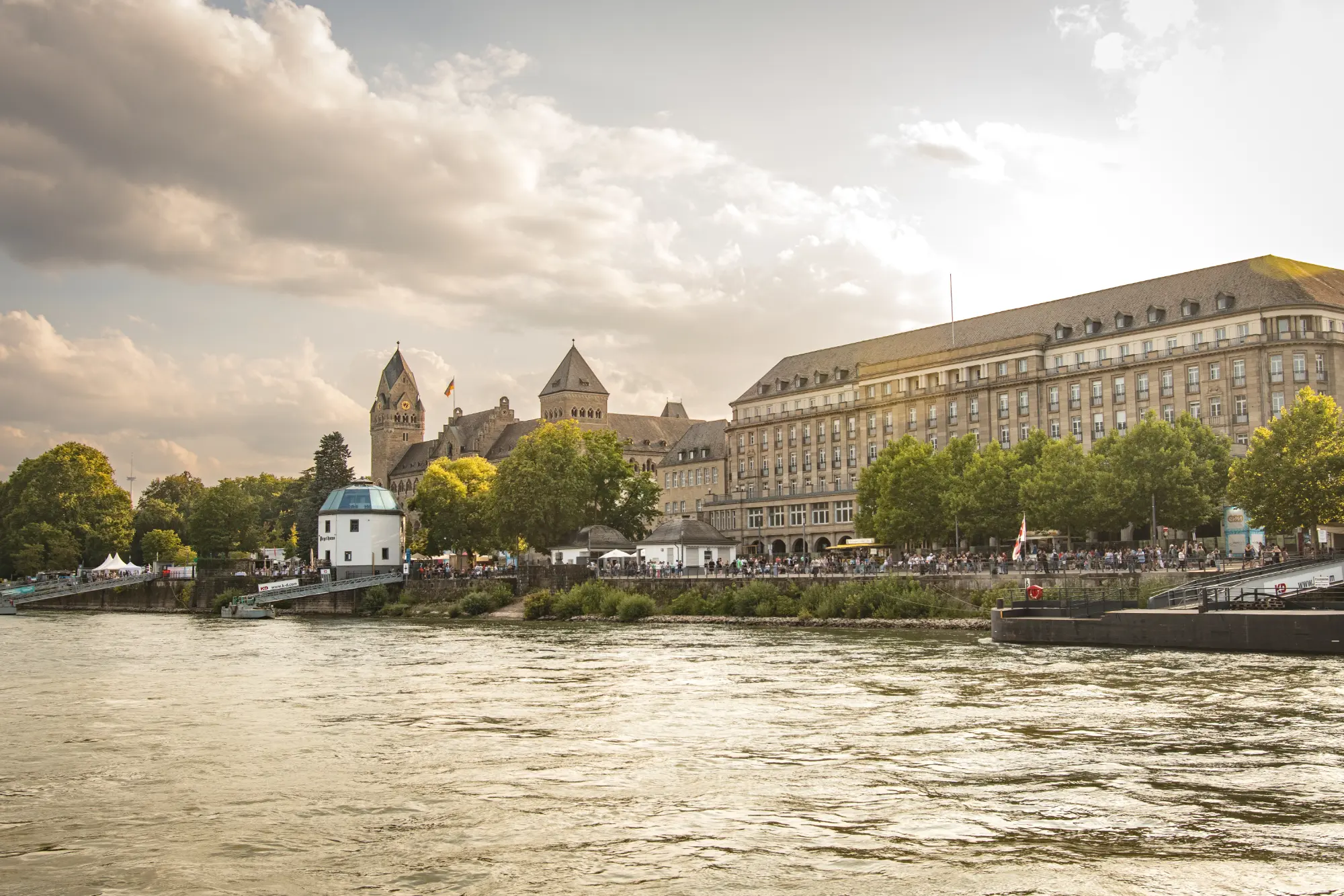 Blick vom Rhein auf das Konrad-Adenauer-Ufer in Koblenz, mit historischen Gebäuden und Bäumen an einem sonnigen Tag.