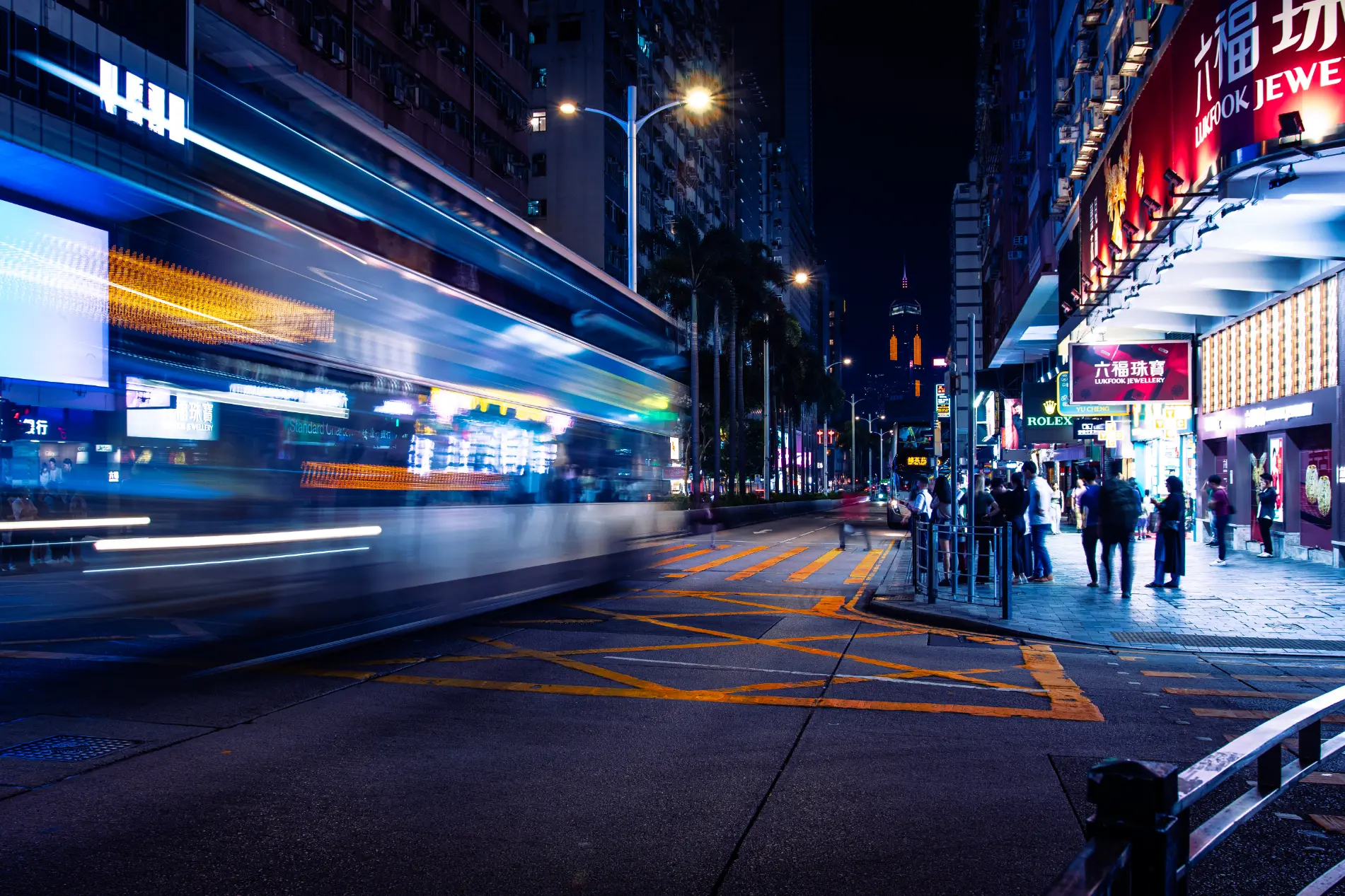 Nächtliche Langzeitbelichtung in einer Straße in Hongkong. Ein vorbeifahrender Doppeldeckerbus wird zu einem blauen Lichtstreifen; die beleuchteten Schilder sind scharf.
