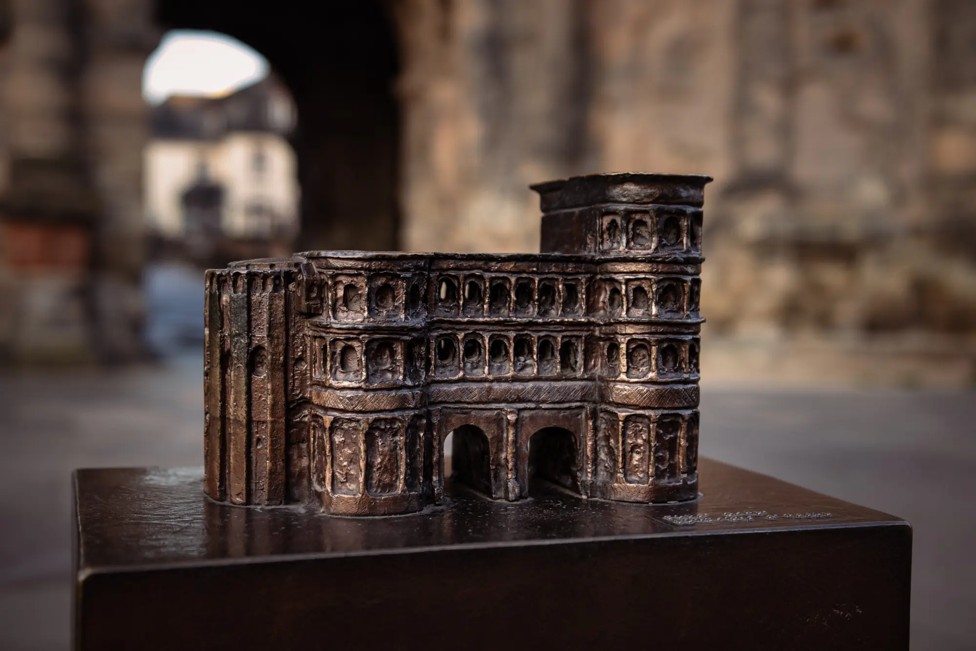 Bronzemodell der Porta Nigra in Trier auf einem Sockel im Vordergrund, mit der echten, unscharfen Porta Nigra im Hintergrund.