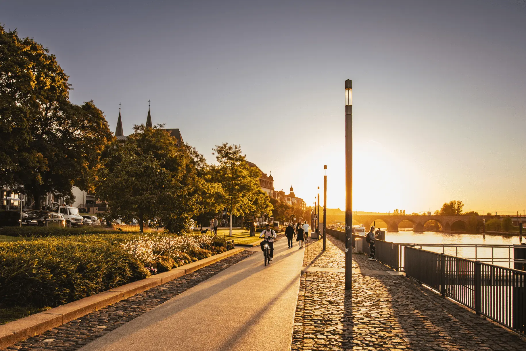 Sonniges Moselufer in Koblenz bei Sonnenuntergang mit einer Promenade. Ein Radfahrer fährt entlang, während die Sonne über der Moselbrücke untergeht.