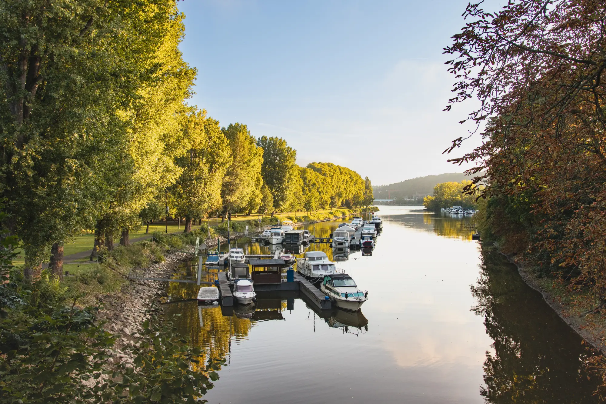 Der Schwanenteich oder ein Hafen in den Kaiserin-Augusta-Anlagen in Koblenz mit am Steg festgemachten kleinen Booten und üppigen Bäumen im Herbstlicht.
