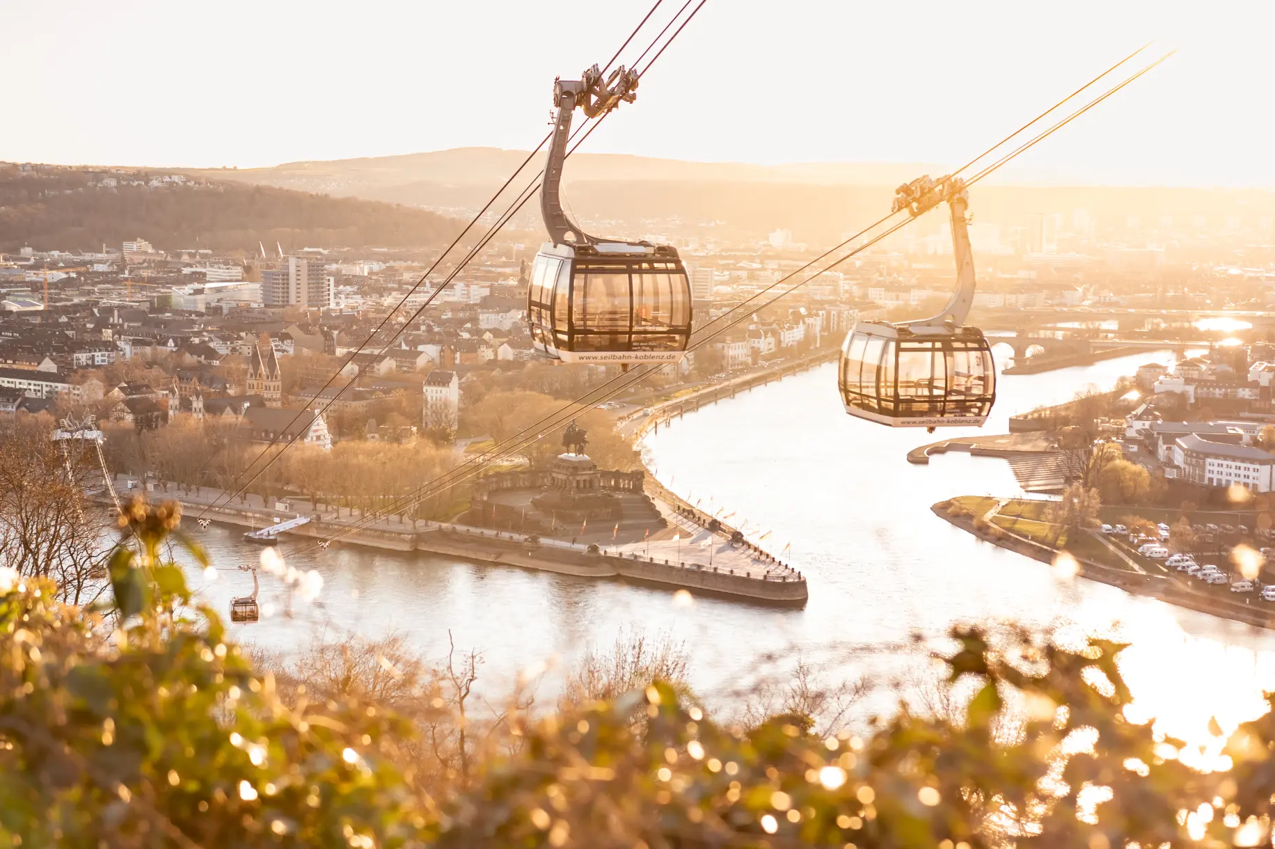 Zwei Seilbahn-Gondeln in der Luft über dem Deutschen Eck in Koblenz bei hellem Sonnenuntergang. Im Hintergrund sieht man den Rhein und die Altstadt.
