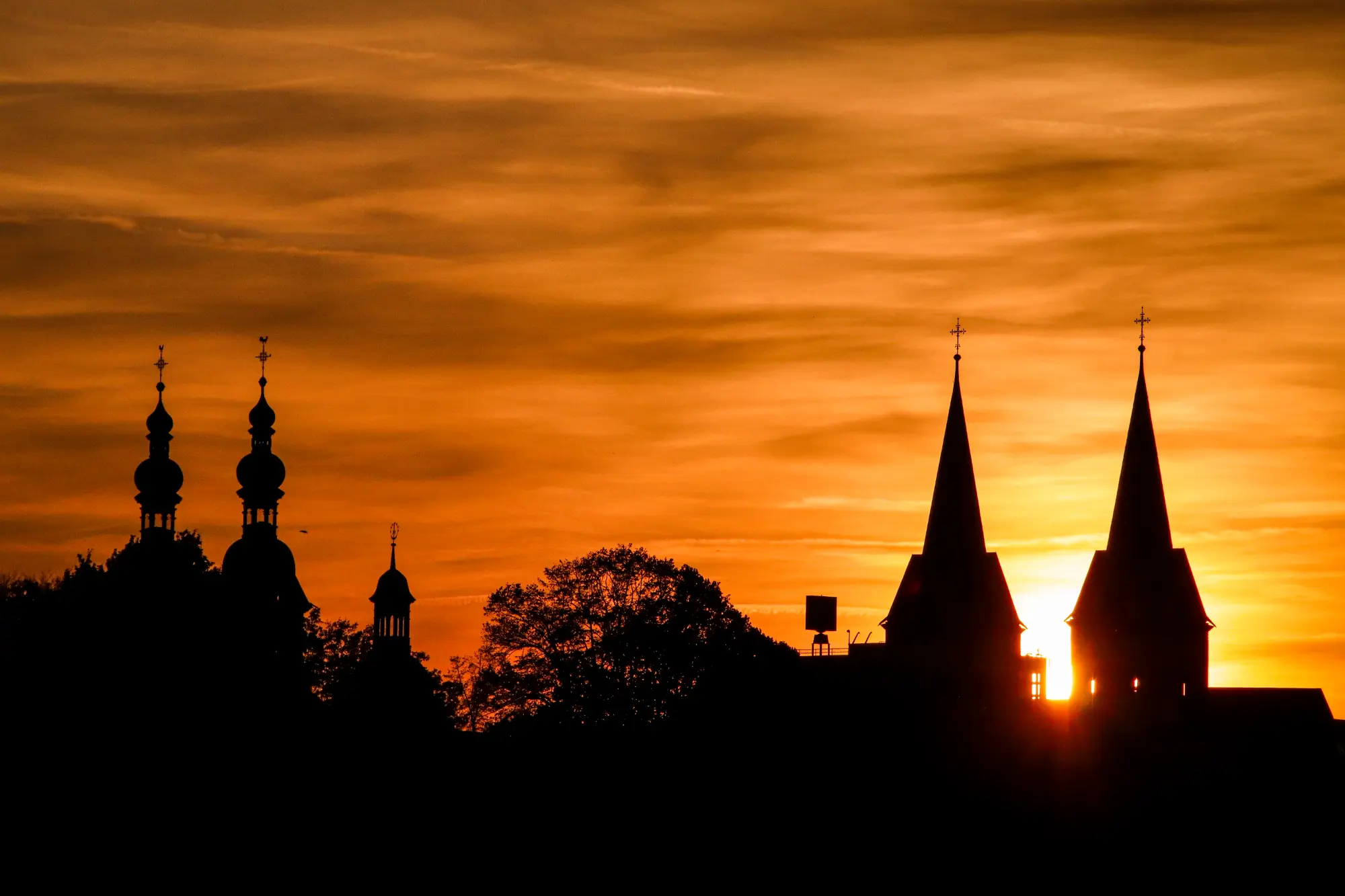 Silhouette der Kirchtürme von Koblenz (vermutlich Liebfrauenkirche und Basilika St. Kastor) vor einem orangefarbenen Sonnenuntergangshimmel.